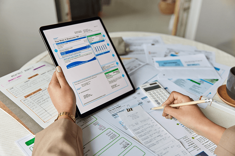 Ręka trzymająca tablet, na którym wyświetlony jest dokument z wykresami, w tle biurko, na nim kalkulator i papierowe wydruki. Ilustracja do wpisu blogowego na temat Jaka stawka VAT na e-booki