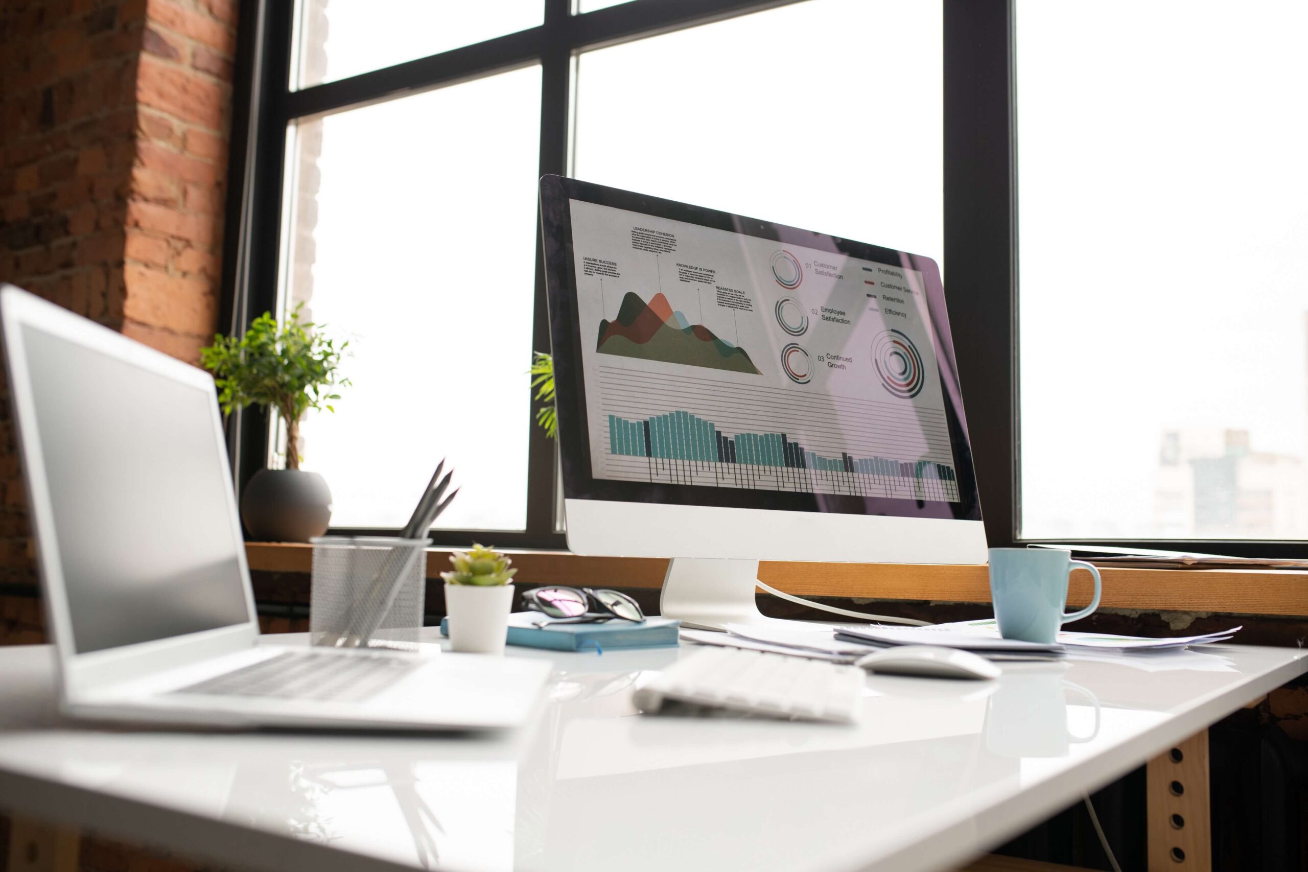 Blat biurka z laptopem, klawiaturą, myszą komputerową, kubkiem i dodatkowym monitorem, na którym widać wykresy. W tle duże okno. Ilustracja do tekstu na temat Jak zaprojektować dobry landing sprzedażowy