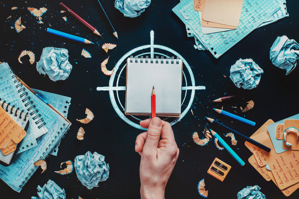 W centralnym punkcie ułożony na narysowanym celowniku notes, nad nim ludzka dłoń z czerwoną kredką wskazującą w środek kartki. Wokół kotesy, zgniecione kartki, kredki i resztki temperowanych ołówków. Ilustracja do wpisu o evergreen content