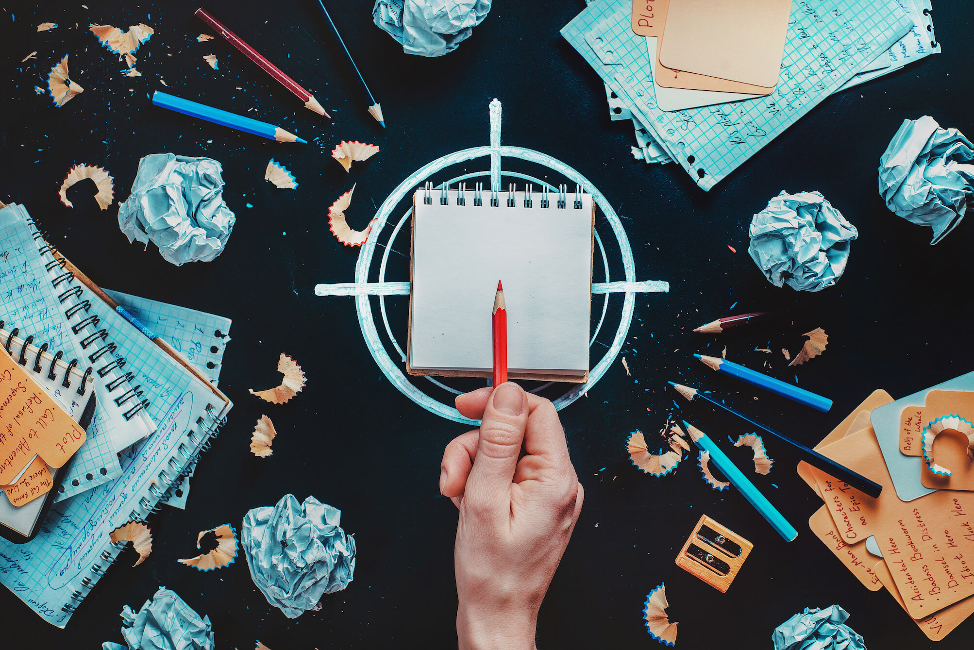 W centralnym punkcie ułożony na narysowanym celowniku notes, nad nim ludzka dłoń z czerwoną kredką wskazującą w środek kartki. Wokół kotesy, zgniecione kartki, kredki i resztki temperowanych ołówków. Ilustracja do wpisu o evergreen content