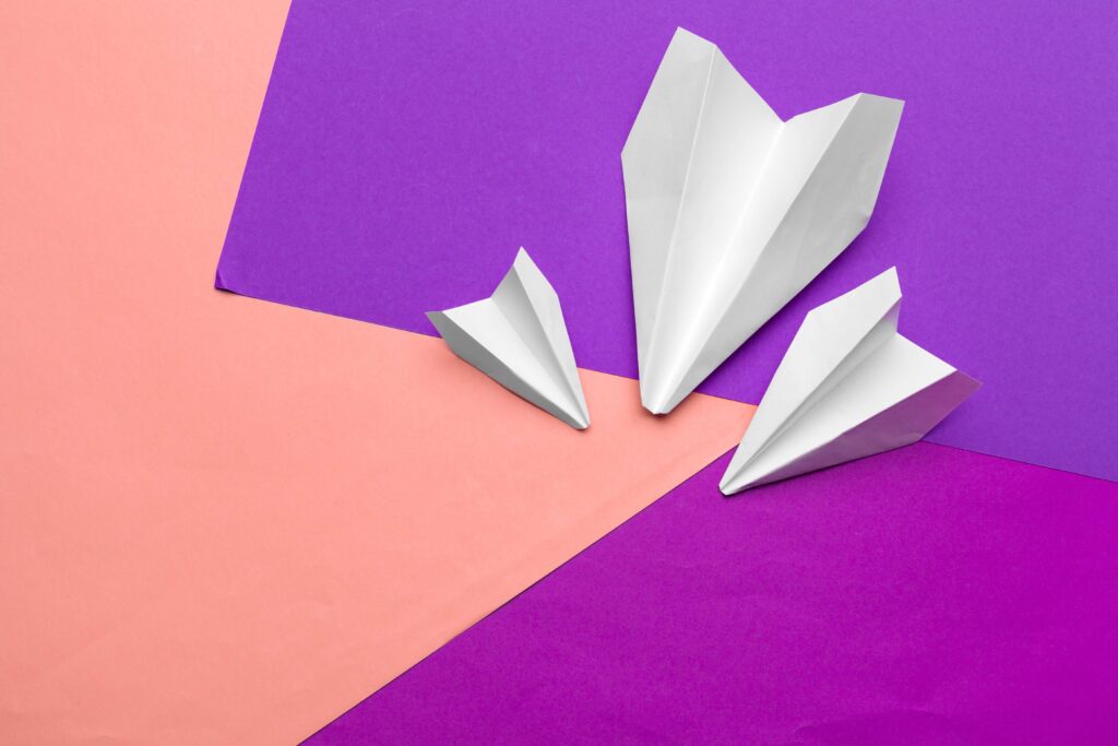 Papierowe samolociki ułożone na różnokolorowym papierze w odcieniach różu i fioletu. Ilustracja do tekstu na temat freebie.