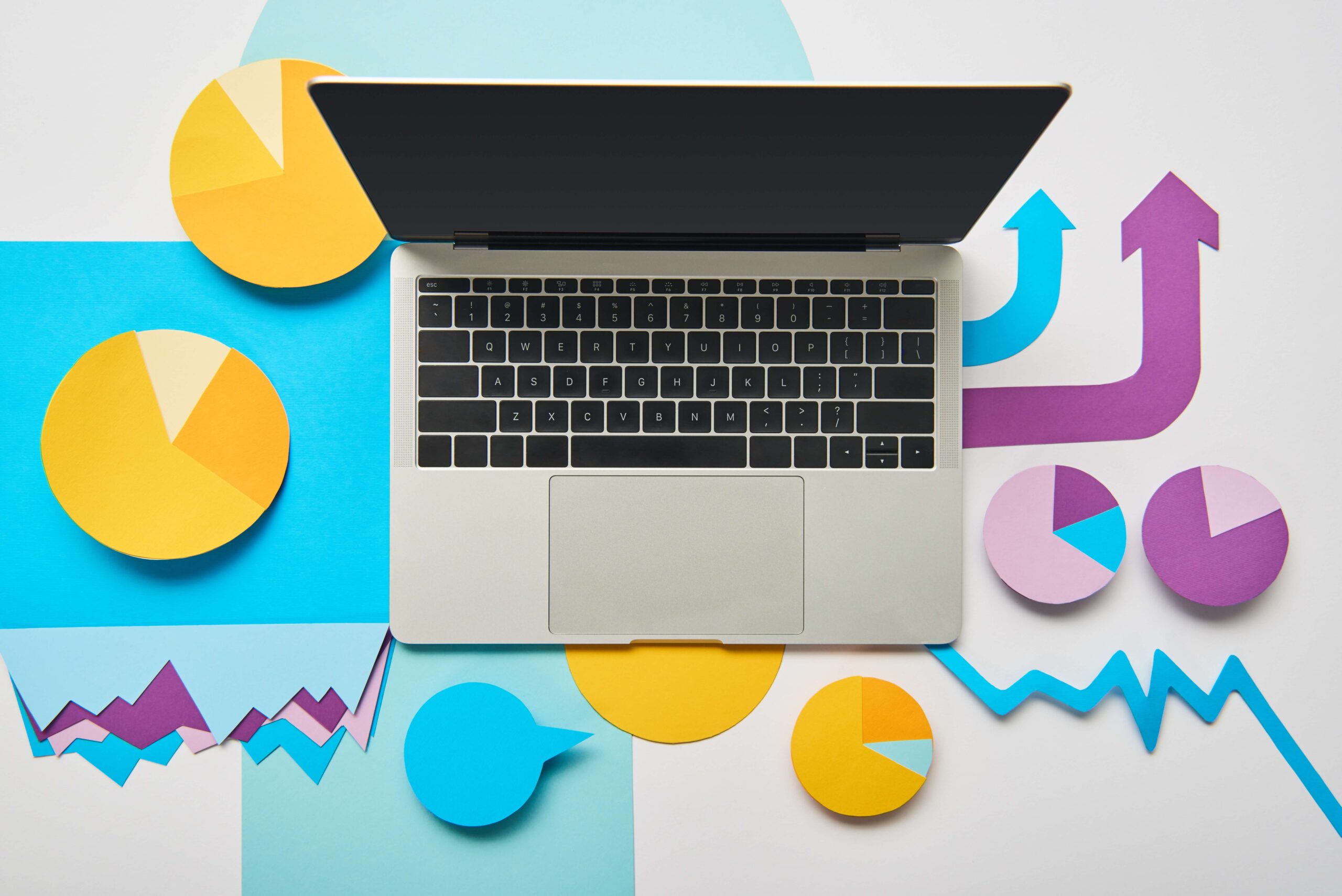 Widziany od góry blat biurka z kolorowymi wyciętymi z papieru wykresami i strzałkami. W centrum otwarty laptop. Ilustracja do tekstu pod tytułem Produkt cyfrowy – rodzaje, koszty, wycena
