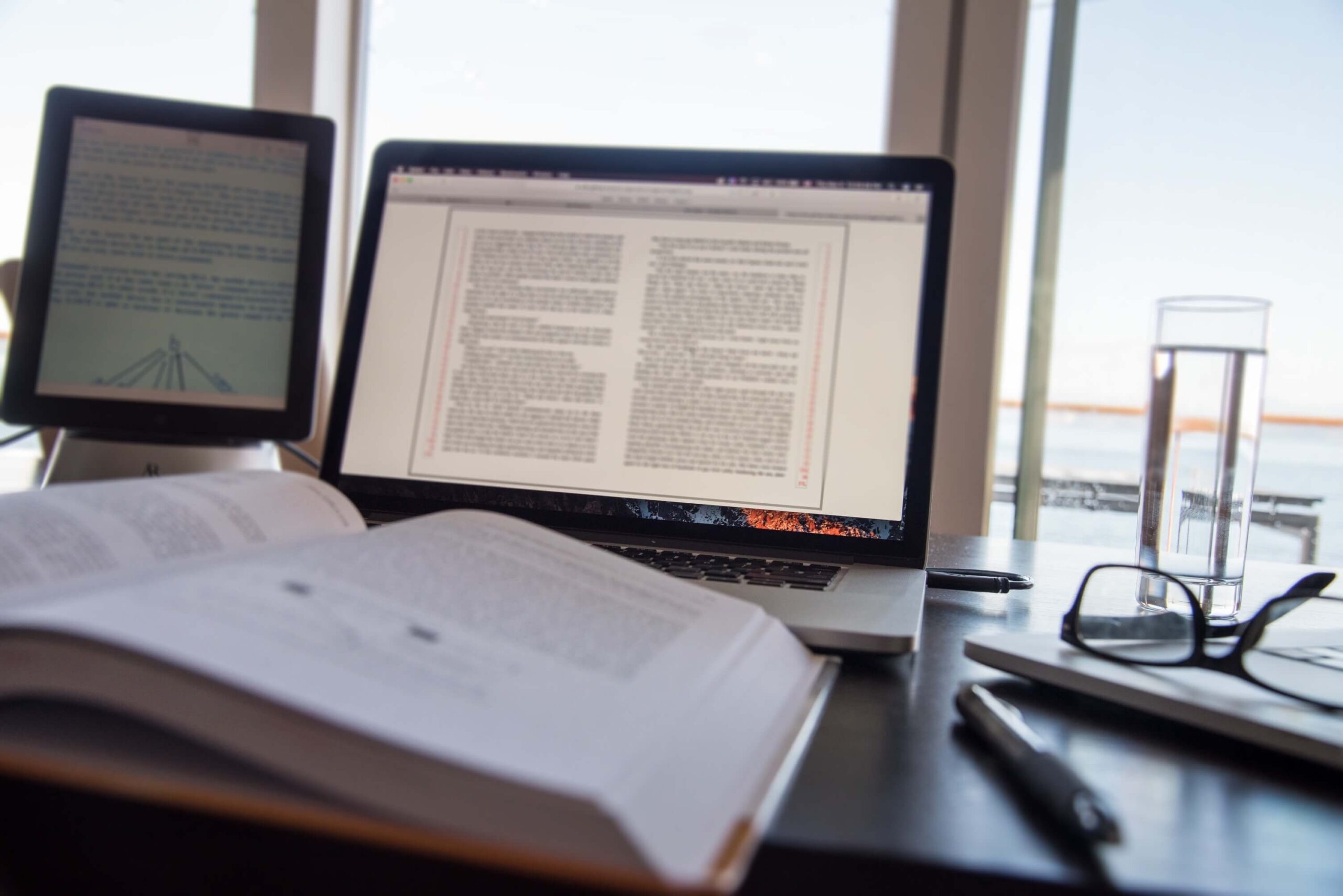 Biurko, a na nim tablet, monitor laptopa, otwarta książka, w tle okulary i szklanka z wodą. Ilustracja do wpisu blogowego pod tytułem Redakcja i korekta e-booka