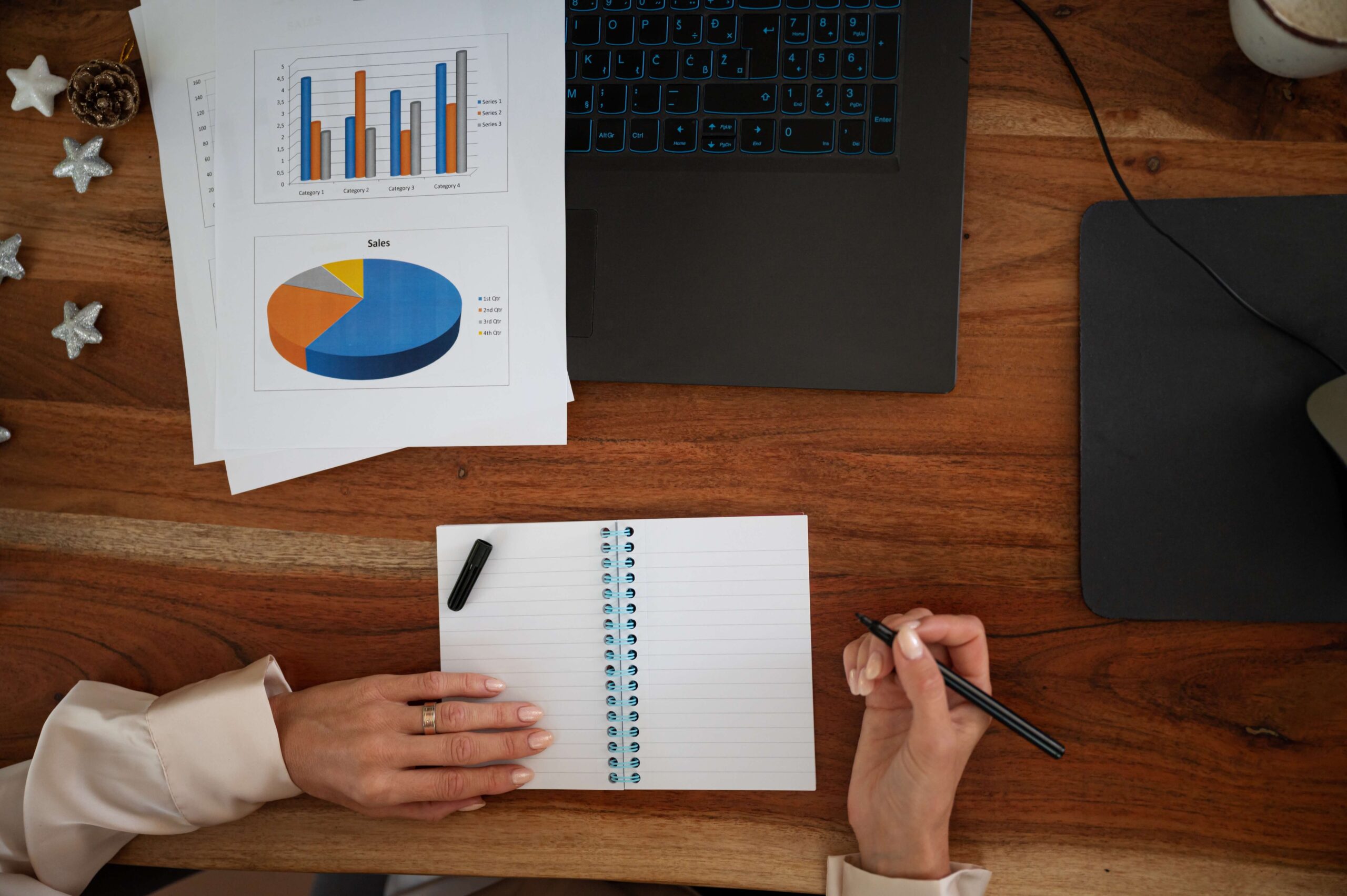 Widok od góry: drewniany blat biurka, kartki z wydrukami, laptop, ręce osoby piszącej w kołozeszycie. Ilustracja do wpisu pod tytułem Project manager w biznesie online