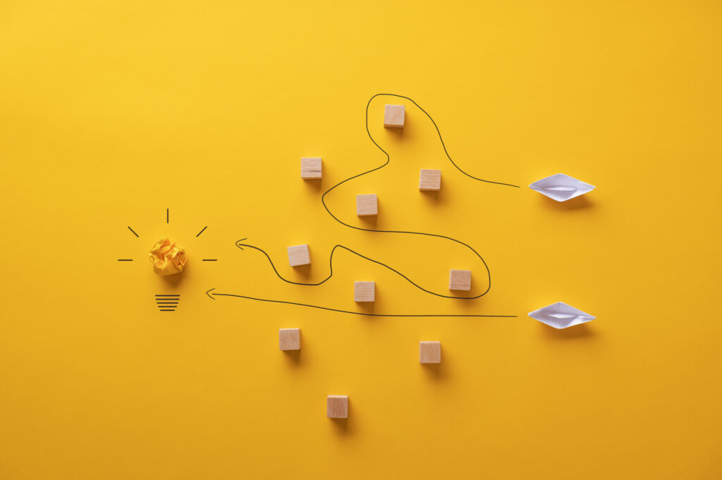 Żółte tło, po lewej stronie zgnieciona żółta kartka imitująca wnętrze żarówki z dorysowanym gwintem i promieniami, w centrum drewniane klocki, po prawej papierowe statki,. Górny ma wytyczoną drogę zygzakiem między klockami, dolny prosto do rozświetlonej żarówki. Ilustracja do tekstu Jak przygotować skuteczny plan contentowy