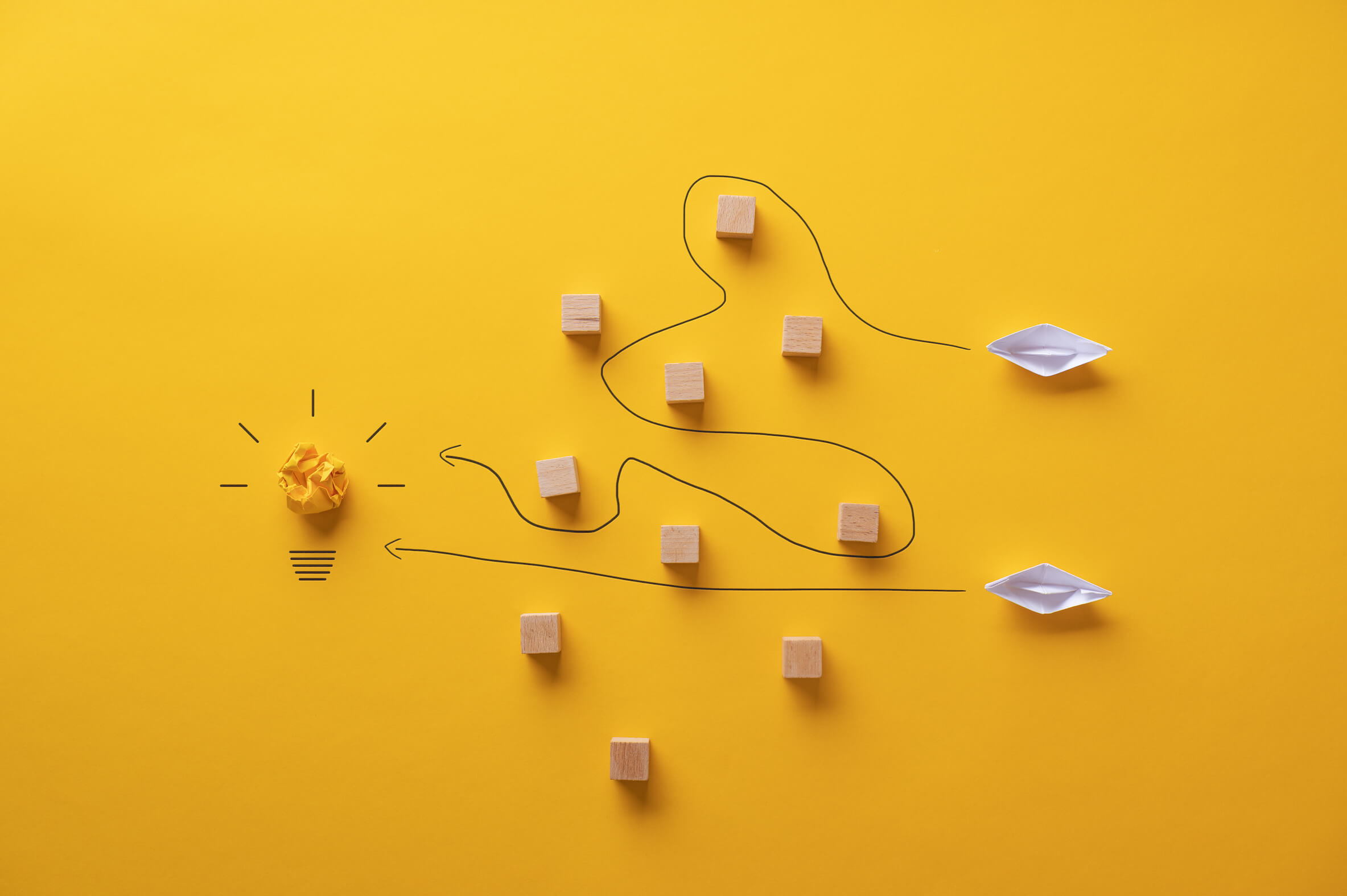 Żółte tło, po lewej stronie zgnieciona żółta kartka imitująca wnętrze żarówki z dorysowanym gwintem i promieniami, w centrum drewniane klocki, po prawej papierowe statki,. Górny ma wytyczoną drogę zygzakiem między klockami, dolny prosto do rozświetlonej żarówki. Ilustracja do tekstu Jak przygotować skuteczny plan contentowy