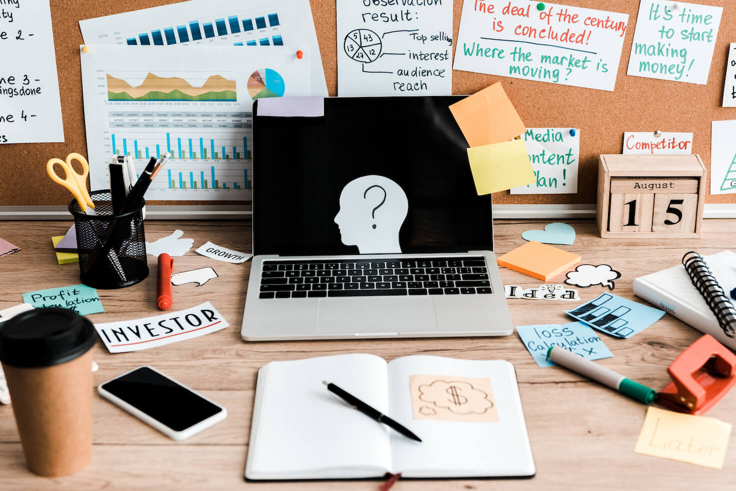 Biurko a na nim w centralnym punkcie laptop z wyciętą z papieru głową i znakiem zapytania, dookoła przybory do pisania, zeszyty, wydruki. Ilustracja do tekstu: Współpraca z copywriterem