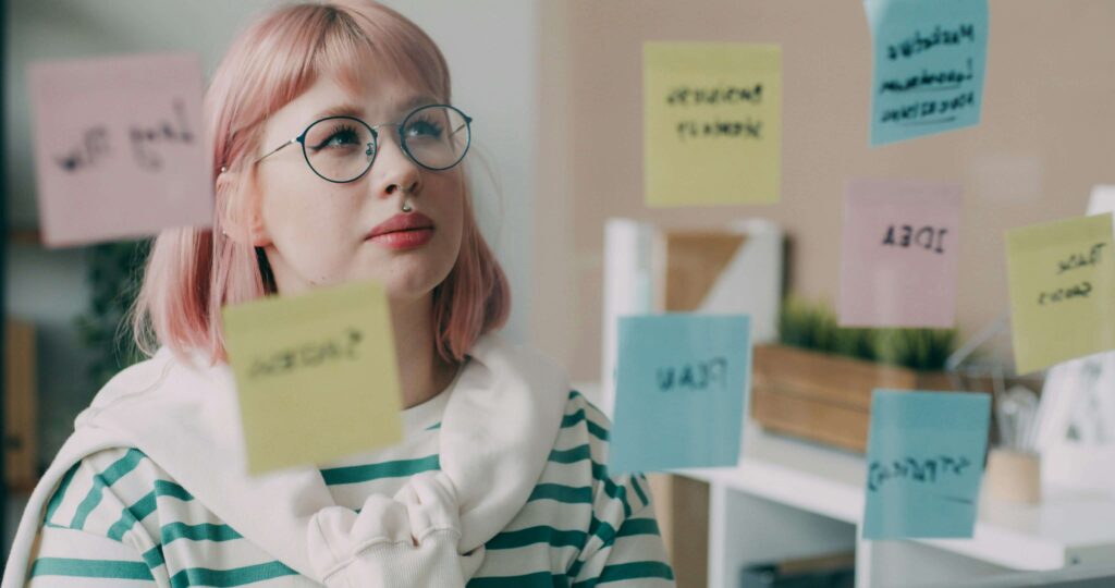 Kobieta w okularach, z jasnymi włosami wpatruje się w szklaną tablicę, do której przyczepione są różnokolorowe karteczki typu post-it z notatkami. Ilustracja do tekstu Persona w biznesie online