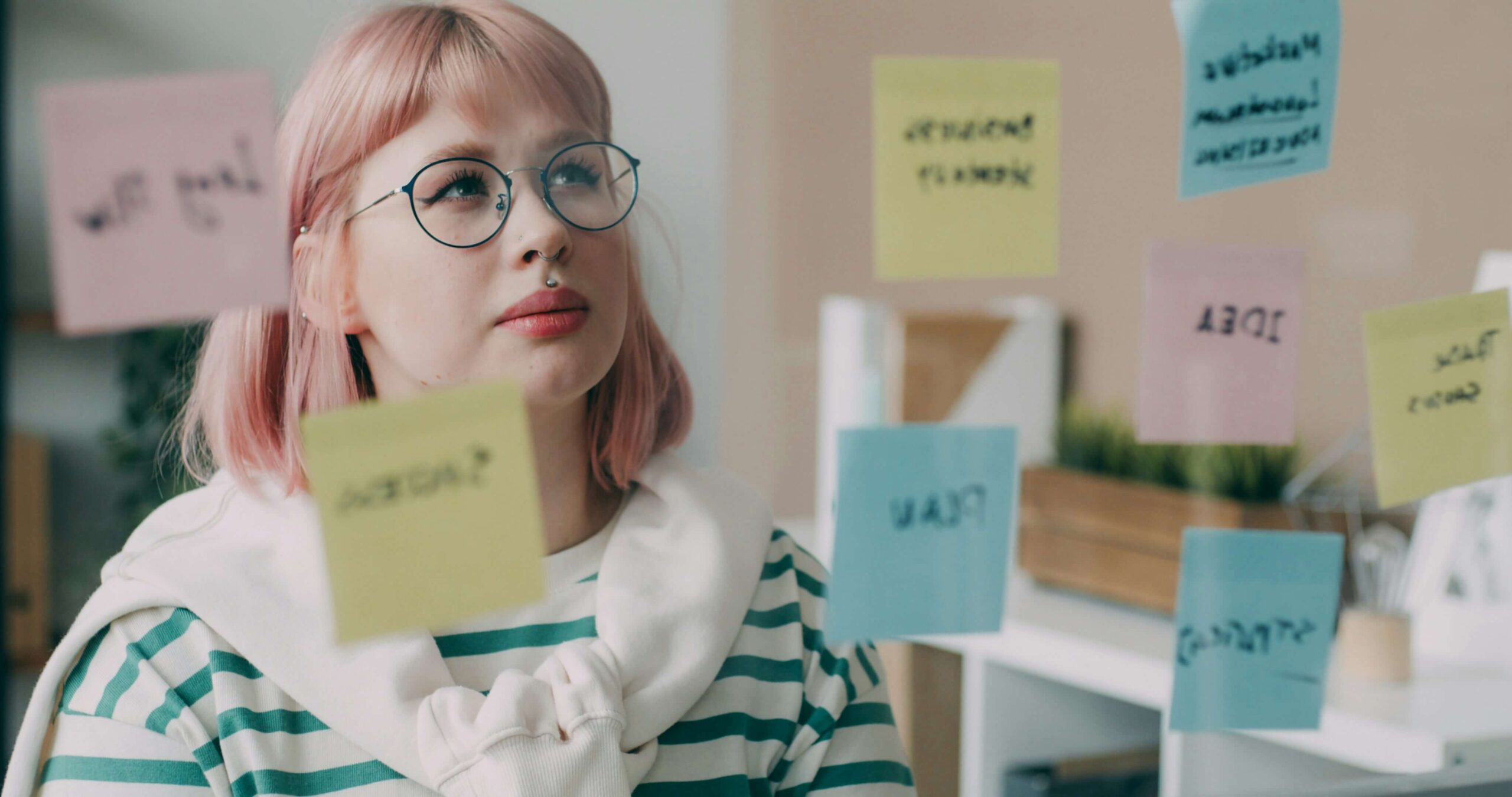 Kobieta w okularach, z jasnymi włosami wpatruje się w szklaną tablicę, do której przyczepione są różnokolorowe karteczki typu post-it z notatkami. Ilustracja do tekstu Persona w biznesie online