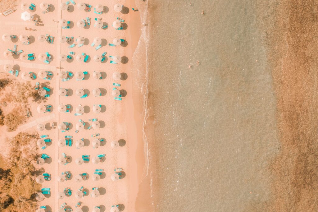 Widok z góry na brzeg morza i piaszczystą plażę, na której w równych odległościach ustawione są rzędy parasoli plażowych. Ilustracja do tekstu Prekampania